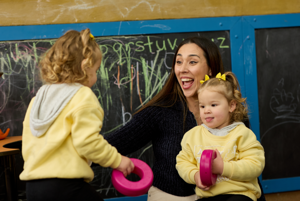 A smiling woman sits with two young children in front of a chalkboard covered in colourful drawings. One child faces her while the other sits beside her holding a pink toy, both wearing yellow tops with matching hair bows. The woman appears engaged and joyful, interacting with them.