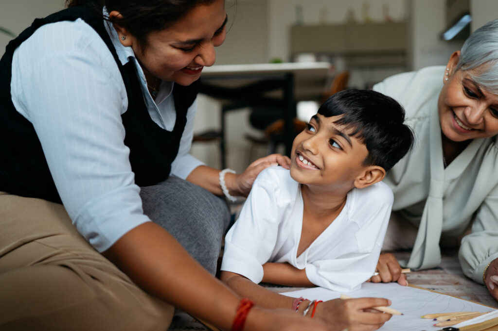 An authentic moment showcasing a grandmother and mother providing gentle encouragement to a young boy as he works on a creative drawing project on the living room floor.